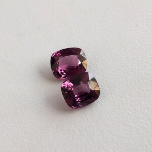 May include: Two cushion-cut gemstones, one on top of the other. The gemstones are a deep purplish-pink color.