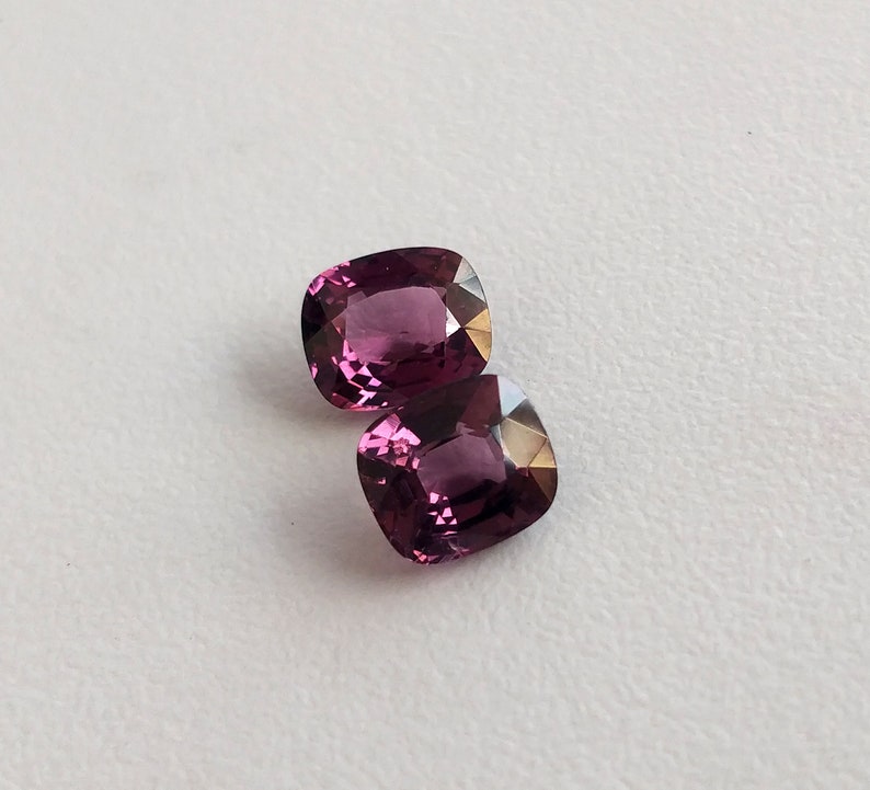 May include: Two cushion-cut gemstones, one on top of the other. The gemstones are a deep purplish-pink color.