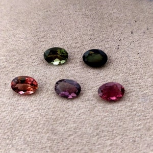 May include: Six oval-shaped gemstones in various shades of green, pink, and purple. The gemstones are arranged in two rows of three on a light brown surface.
