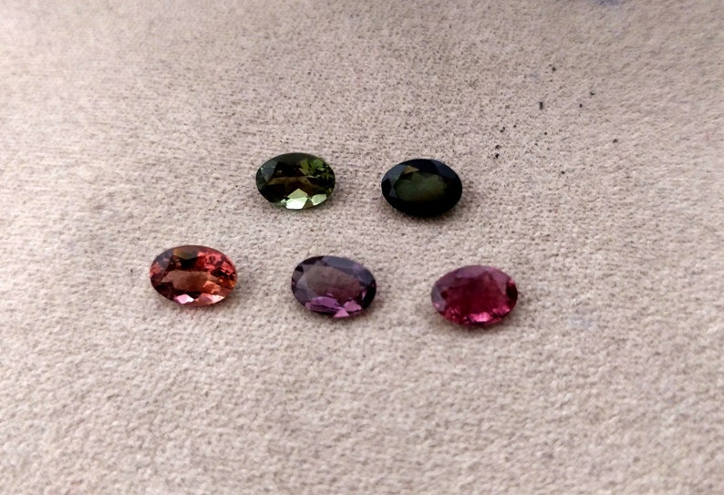 May include: Six oval-shaped gemstones in various shades of green, pink, and purple. The gemstones are arranged in two rows of three on a light brown surface.