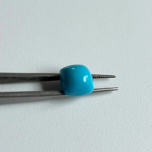 May include: A turquoise blue bead held in silver tweezers. The bead is round and smooth.