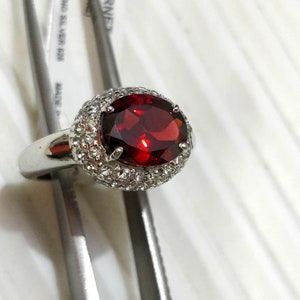 May include: A silver ring with a large red oval gemstone surrounded by a halo of smaller white gemstones.