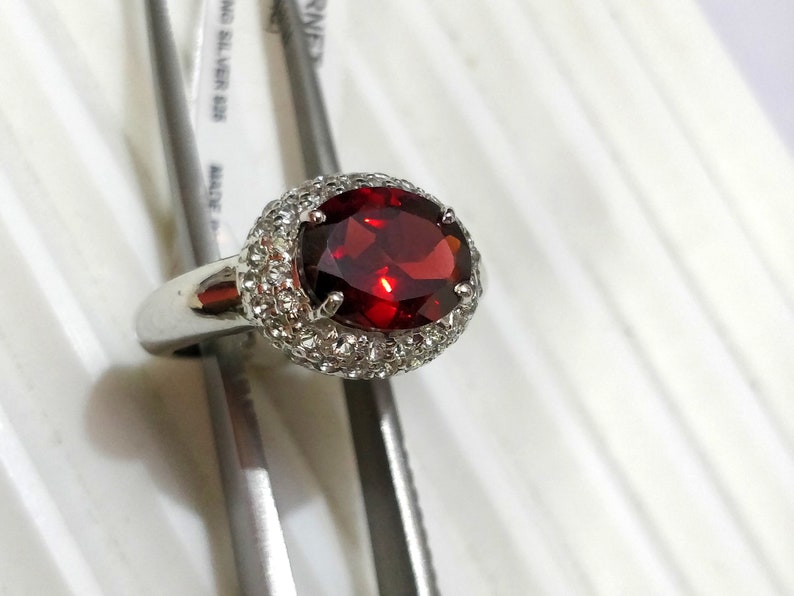 May include: A silver ring with a large red oval gemstone surrounded by a halo of smaller white gemstones.