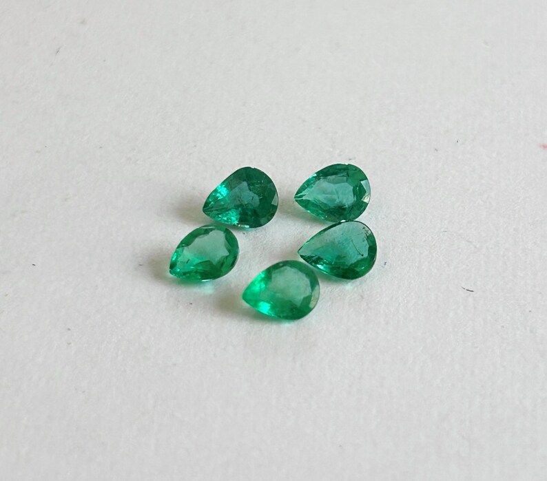 K&ouml;nnte beinhalten: F&uuml;nf lose, birnenf&ouml;rmige Smaragd-Edelsteine mit leuchtend gr&uuml;ner Farbe und facettierten Oberfl&auml;chen. Jeder Stein weist eine klare, durchscheinende Qualit&auml;t mit nat&uuml;rlichen Einschl&uuml;ssen auf und ist in einer Gruppe auf einer hellen Fl&auml;che angeordnet.