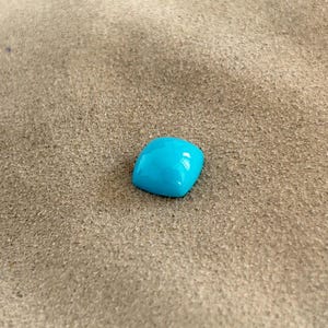 May include: A bright turquoise gemstone, likely turquoise, is displayed on a light brown, textured surface.