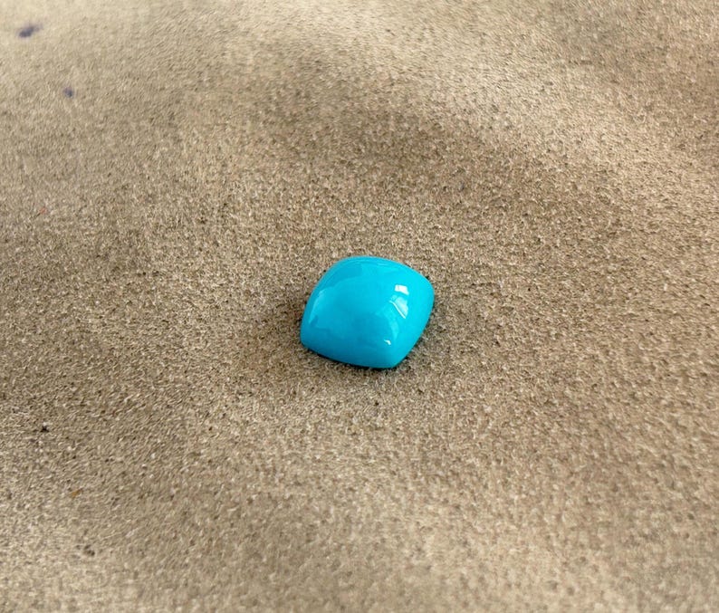 May include: A bright turquoise gemstone, likely turquoise, is displayed on a light brown, textured surface.