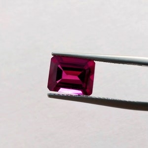 May include: A deep pink, emerald-cut gemstone held in tweezers. The gemstone is faceted and has a high shine.