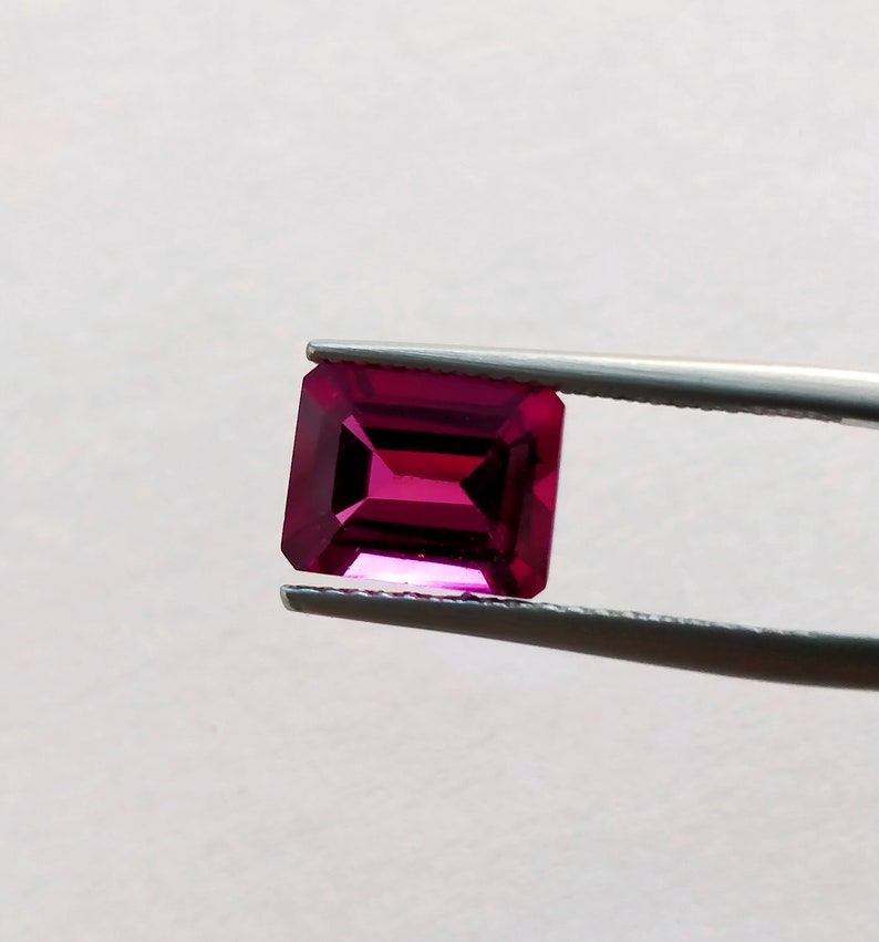May include: A deep pink, emerald-cut gemstone held in tweezers. The gemstone is faceted and has a high shine.