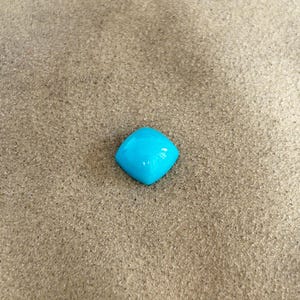 May include: A bright blue, square-shaped gemstone with a smooth, polished surface. The gemstone is sitting on a light brown, sandy surface.