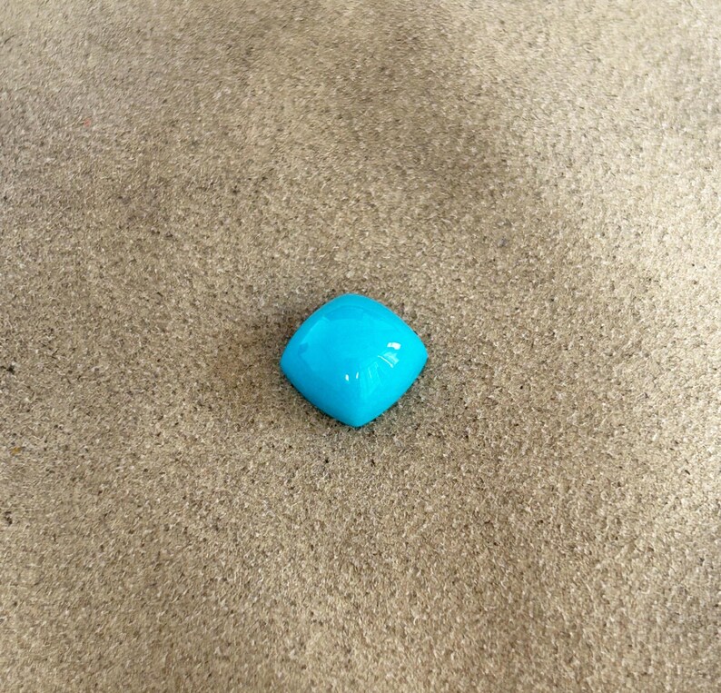 May include: A bright blue, square-shaped gemstone with a smooth, polished surface. The gemstone is sitting on a light brown, sandy surface.