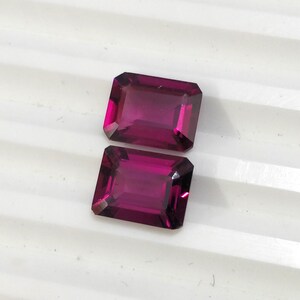 May include: Two rectangular, faceted gemstones with a deep purple hue. The gemstones are cut in an emerald shape.