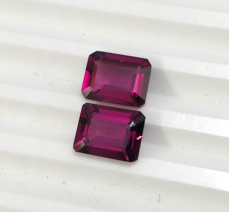 May include: Two rectangular, faceted gemstones with a deep purple hue. The gemstones are cut in an emerald shape.