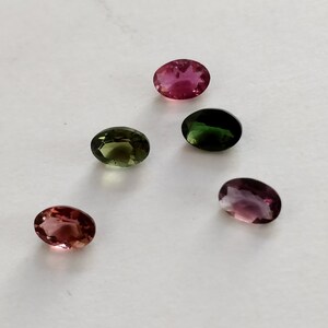 May include: Five oval-shaped gemstones in various shades of pink, green, and purple. The gemstones are arranged in a semi-circle on a white surface.