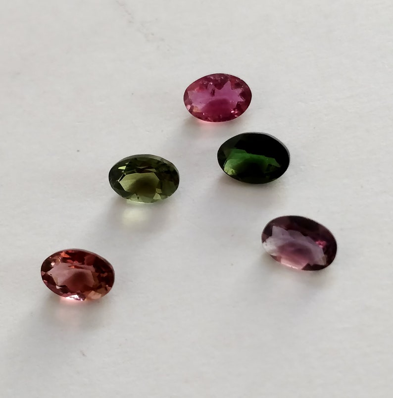 May include: Five oval-shaped gemstones in various shades of pink, green, and purple. The gemstones are arranged in a semi-circle on a white surface.