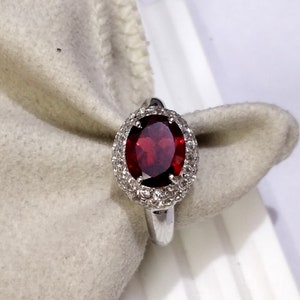 May include: A silver ring with a large oval-shaped red gemstone surrounded by a halo of smaller white gemstones.
