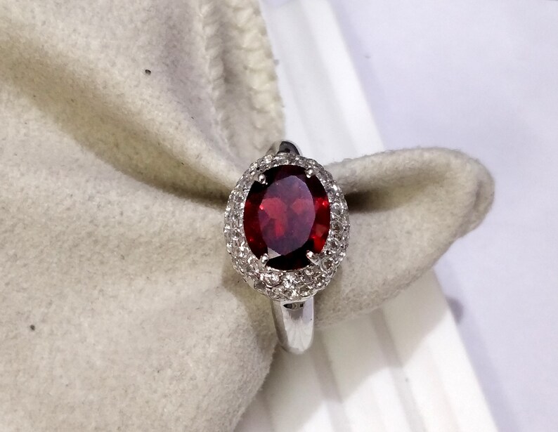 May include: A silver ring with a large oval-shaped red gemstone surrounded by a halo of smaller white gemstones.