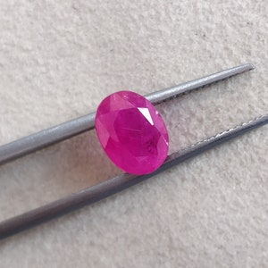 Puede incluir: Una gema ovalada de color rosa vibrante sostenida por unas pinzas. La gema tiene un corte facetado y es de color rosa intenso.