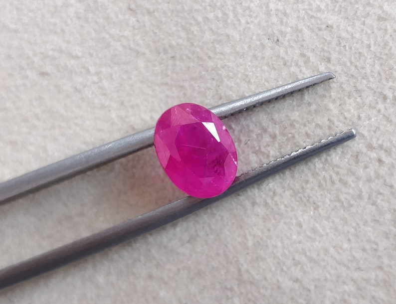 Puede incluir: Una gema ovalada de color rosa vibrante sostenida por unas pinzas. La gema tiene un corte facetado y es de color rosa intenso.