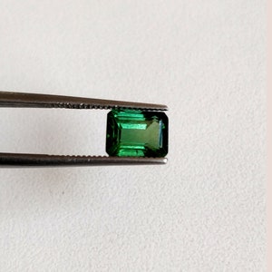 May include: A vibrant green emerald gemstone, cut in a rectangular emerald shape, is held by tweezers against a white background.