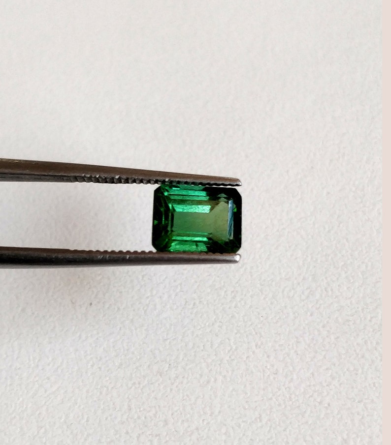 May include: A vibrant green emerald gemstone, cut in a rectangular emerald shape, is held by tweezers against a white background.