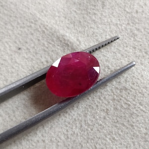 Puede incluir: Una gema ovalada de color rojo vibrante sostenida por unas pinzas. La gema es lisa y pulida, con un tono rojo intenso.