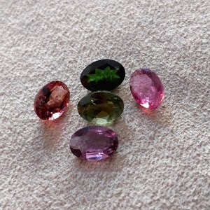 May include: Five oval-shaped gemstones in various colors, including pink, green, and purple. The gemstones are arranged in a semi-circle on a white surface.