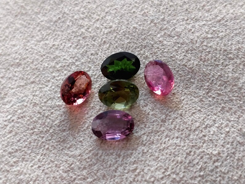 May include: Five oval-shaped gemstones in various colors, including pink, green, and purple. The gemstones are arranged in a semi-circle on a white surface.
