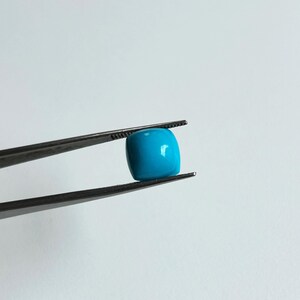 May include: A bright blue, square-shaped turquoise gemstone held in tweezers.