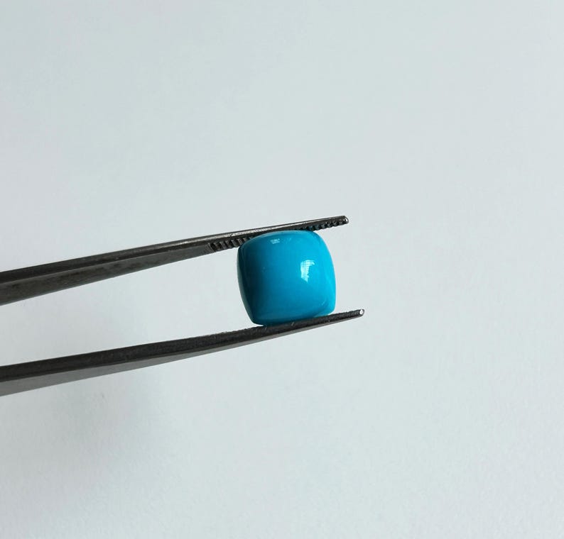 May include: A bright blue, square-shaped turquoise gemstone held in tweezers.