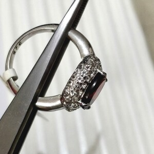 May include: A silver ring with a large, oval-shaped, dark red gemstone surrounded by smaller, clear gemstones.