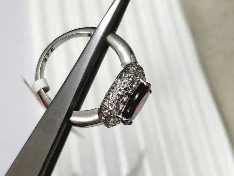 May include: A silver ring with a large, oval-shaped, dark red gemstone surrounded by smaller, clear gemstones.