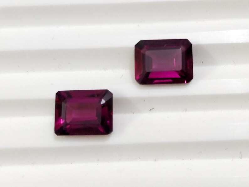 May include: Two purple rectangular gemstones with a faceted cut.