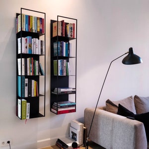 Può includere: Due librerie in metallo nero montate su una parete bianca, ciascuna piena di libri. Le librerie sono in orientamento verticale e hanno ciascuna quattro ripiani. Una lampada da terra nera con un paralume bianco è in primo piano.