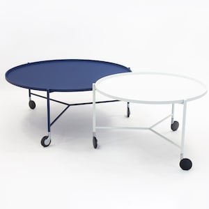 Könnte beinhalten: Zwei runde, rollbare Tische, einer blau und einer weiß. Die Tische haben ein schlichtes, modernes Design mit einer runden Tischplatte und einem dreibeinigen Gestell.