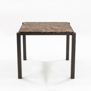 Peut inclure: Une table carrée avec un plateau en marbre brun et un piétement en métal noir. La table a quatre pieds et est sur un fond blanc.