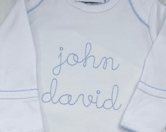 May include: A white long-sleeve baby gown with light blue trim along the neckline and cuffs. The name John David is embroidered in light blue cursive script across the chest. A white tag at the neck reads Bayou Blanks and indicates a 0-6m size.