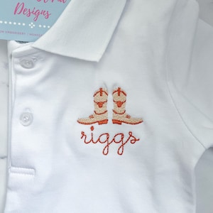 K&ouml;nnte beinhalten: Wei&szlig;es Poloshirt mit orangefarbenen gestickten Cowboystiefeln und dem Namen "riggs" darunter.
