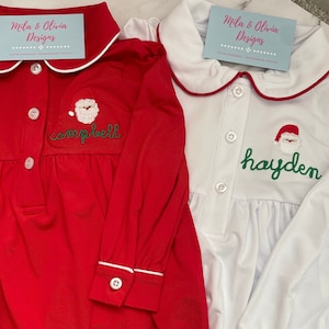 May include: Two red and white Christmas pajamas with Santa Claus embroidery. The red pajamas have the name "campbell" embroidered on the front. The white pajamas have the name "hayden" embroidered on the front.
