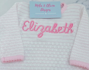 Puede incluir: Un pelele de bebé a rayas blancas y rosas con el nombre "Elizabeth" bordado en rosa en la parte delantera. El pelele tiene un ribete rosa alrededor del cuello y las mangas.