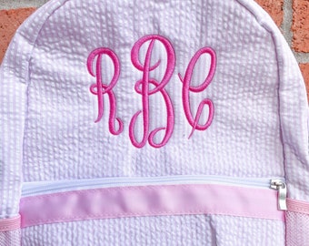 May include: A light pink and white seersucker backpack featuring a front zippered pocket with a horizontal pink ribbon trim. The center of the backpack is embroidered with the large pink monogram letters RBC in a cursive script font.