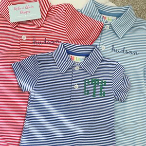 May include: Three short-sleeved polo rompers featuring horizontal stripes in red, light blue, and dark blue. Each garment has a collar, a button placket, and snap closures at the bottom. The red and light blue rompers feature the embroidered name hudson.