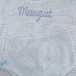 May include: A white baby outfit with light blue stripes and trim. The outfit includes a top with the name "Margot" embroidered in blue and pink, and matching shorts with a small blue bow.