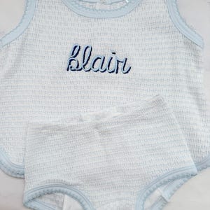 May include: A baby outfit set in white and light blue, featuring a sleeveless top and matching shorts. The top is embroidered with the name "Blair" in navy blue script. The edges are trimmed with a scalloped light blue border.