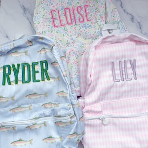 May include: Three personalized backpacks. One is floral with "Eloise" embroidered in pink. Another is light blue with fish and "Ryder" in green. The third is pink gingham with "Lily" in lavender.