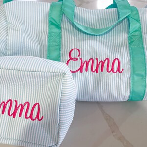 May include: A light blue and white pinstriped duffel bag and a matching rectangular toiletry bag, both featuring the name Emma embroidered in bright pink cursive script. The bags have vibrant teal fabric handles and trim accents.
