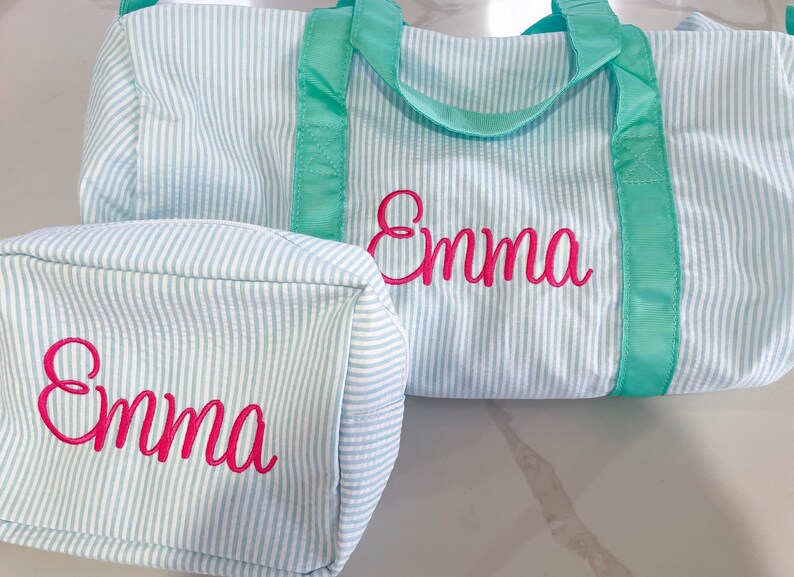 May include: A light blue and white pinstriped duffel bag and a matching rectangular toiletry bag, both featuring the name Emma embroidered in bright pink cursive script. The bags have vibrant teal fabric handles and trim accents.