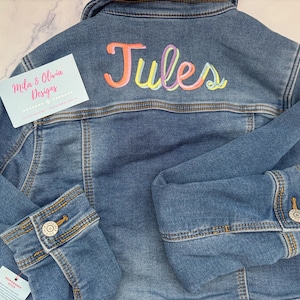 May include: A blue denim jacket with the name "Jules" embroidered on the back in colorful thread. The jacket has buttoned cuffs and a collar. A tag that says "Mila & Olivia Designs" is attached.