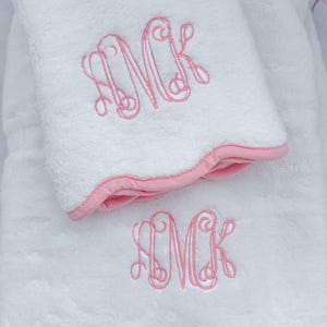 May include: Two white terry cloth towels with pink trim and embroidered pink initials. The towels have a scalloped edge and a soft, plush appearance. The embroidered initials are in a decorative script font.