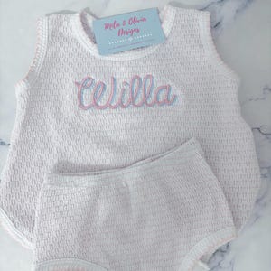 May include: White sleeveless baby onesie with pink and blue embroidered lettering that says "Celilla" and a matching white diaper cover with pink trim.