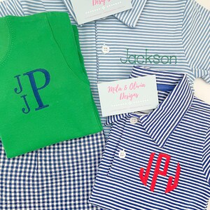 May include: A green t-shirt with a dark blue embroidered monogram, a light blue and white striped polo shirt with the green embroidered name Jackson, and a navy and white striped polo shirt with a red embroidered monogram, plus two business cards.